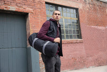 Load image into Gallery viewer, Male model standing with Black duffel bag with black and white checkered shoulder strap
